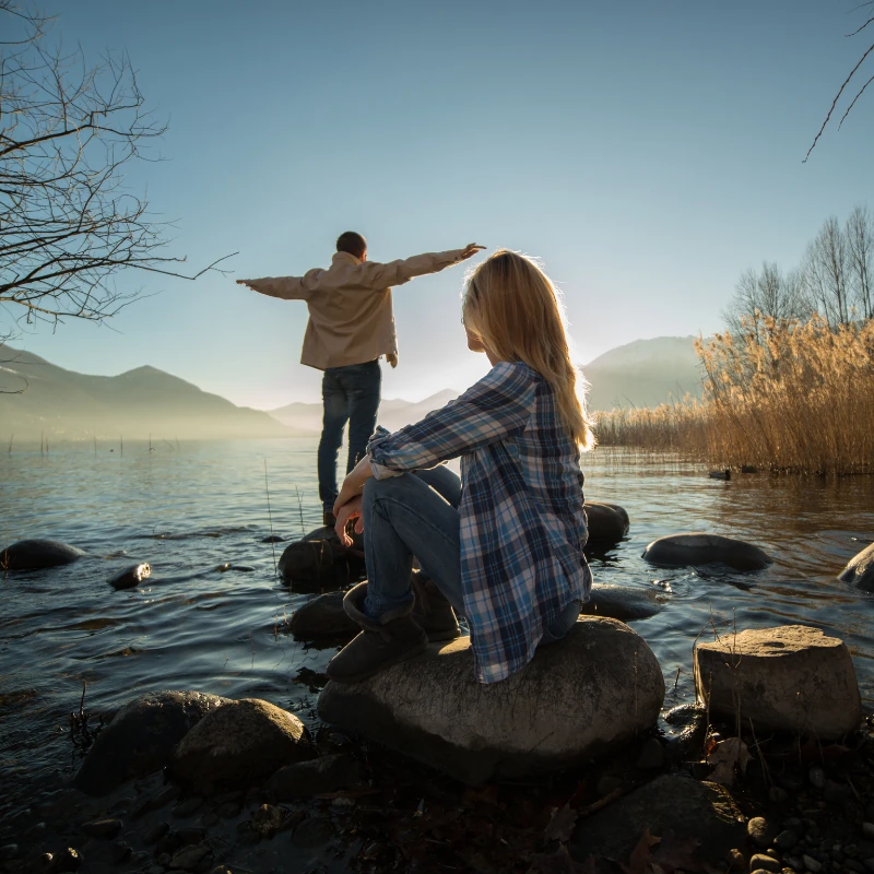 Eine Frau sitzt auf einem Stein am Ufer eines Sees und blickt auf eine Person, die mit ausgebreiteten Armen über Steine im flachen Wasser balanciert; im Hintergrund sind Berge und warmes Abendlicht zu sehen.