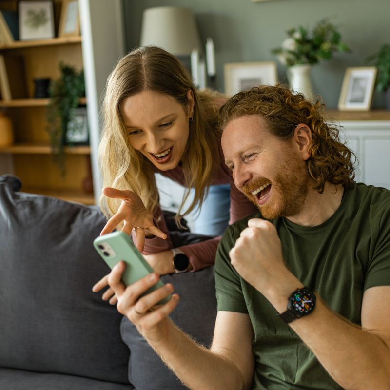 Zwei Personen sitzen auf einem Sofa, schauen gemeinsam auf ein Smartphone und reagieren begeistert und lachend auf den Inhalt.