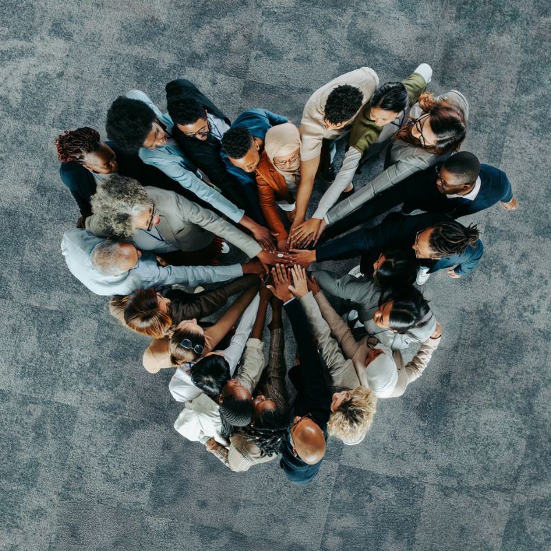 Eine Gruppe von Menschen steht im Kreis und legt die Hände in der Mitte übereinander als Zeichen für Zusammenhalt und Teamarbeit.
